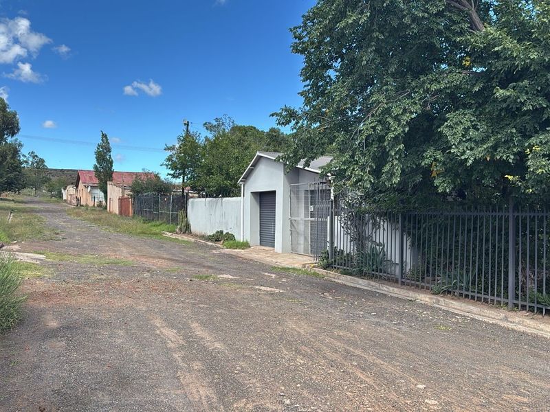 Street view showing the property exterior, featuring metal perimeter fencing, a garage entrance, and boundary walling along an unpaved road.