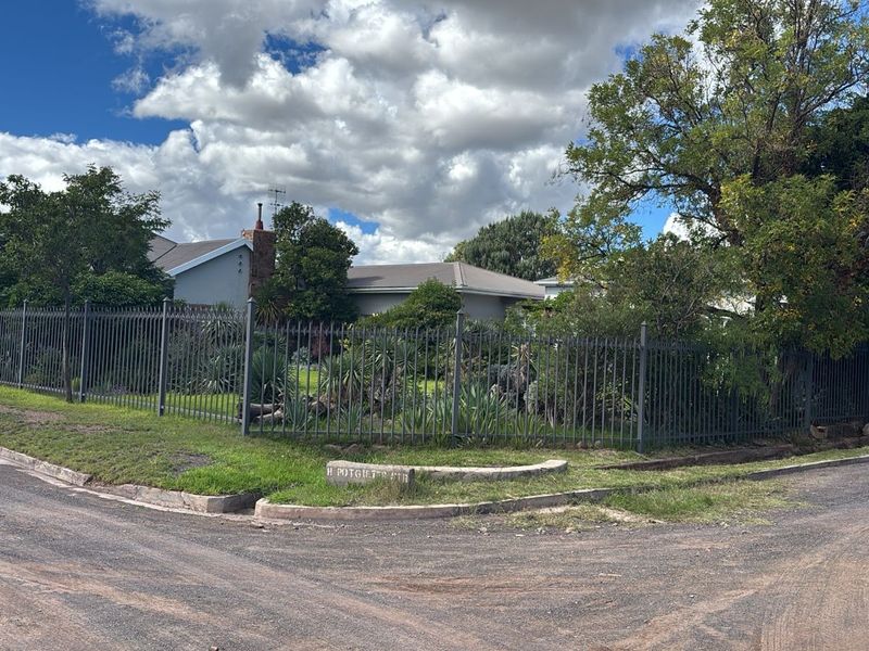 A street-level view of the property featuring a secure metal perimeter fence, established garden, and tiled roof structure.