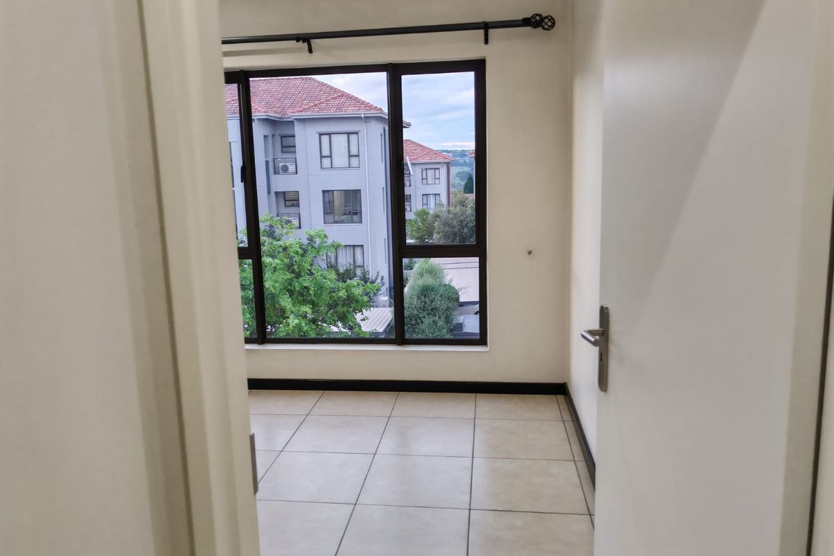 An empty bedroom featuring neutral tiled flooring, a large window with a curtain rod, and views of the surrounding apartment complex.