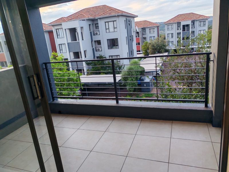 A clean, tiled balcony featuring a modern metal balustrade and sliding glass door access, overlooking the apartment complex grounds.