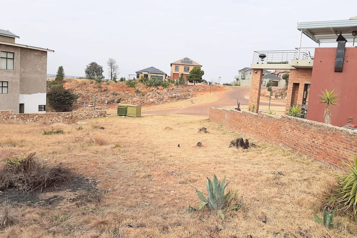 A view of a vacant residential plot featuring dry grass, a brick boundary wall, and surrounding neighborhood homes.