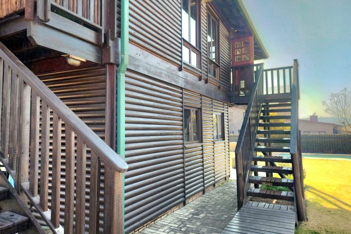Rear view of a timber log cabin featuring an external wooden staircase, balcony, and paved walkway leading to the garden area.