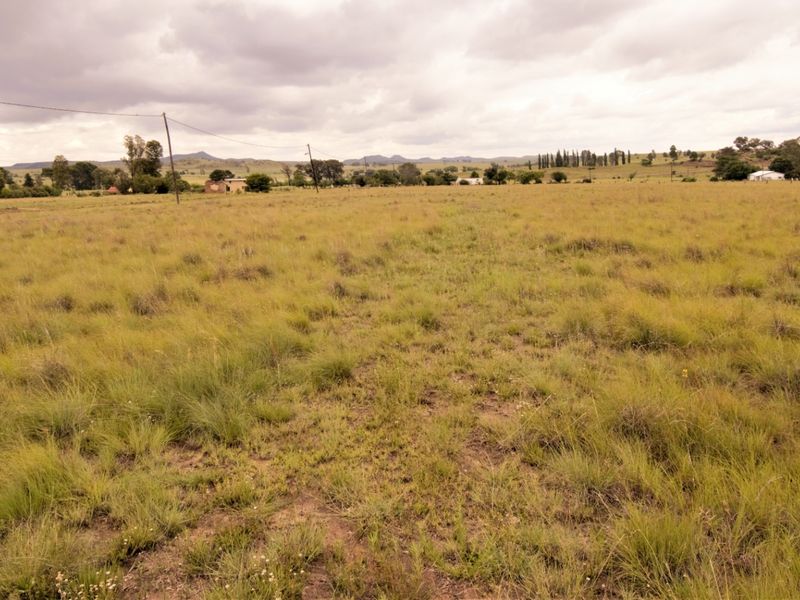 A wide-angle view of a vacant residential plot featuring open grassland and distant rural landscape under a cloudy sky.