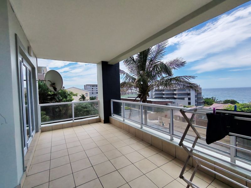 A spacious covered balcony featuring tiled flooring, a modern glass balustrade, and expansive views of the surrounding area and ocean.