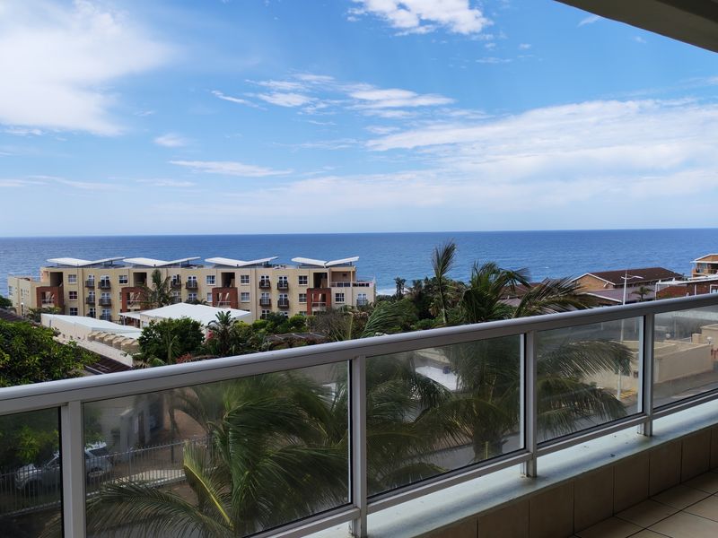 A clear elevated view of the ocean and surrounding coastal apartment complex from a private balcony with a modern glass balustrade.