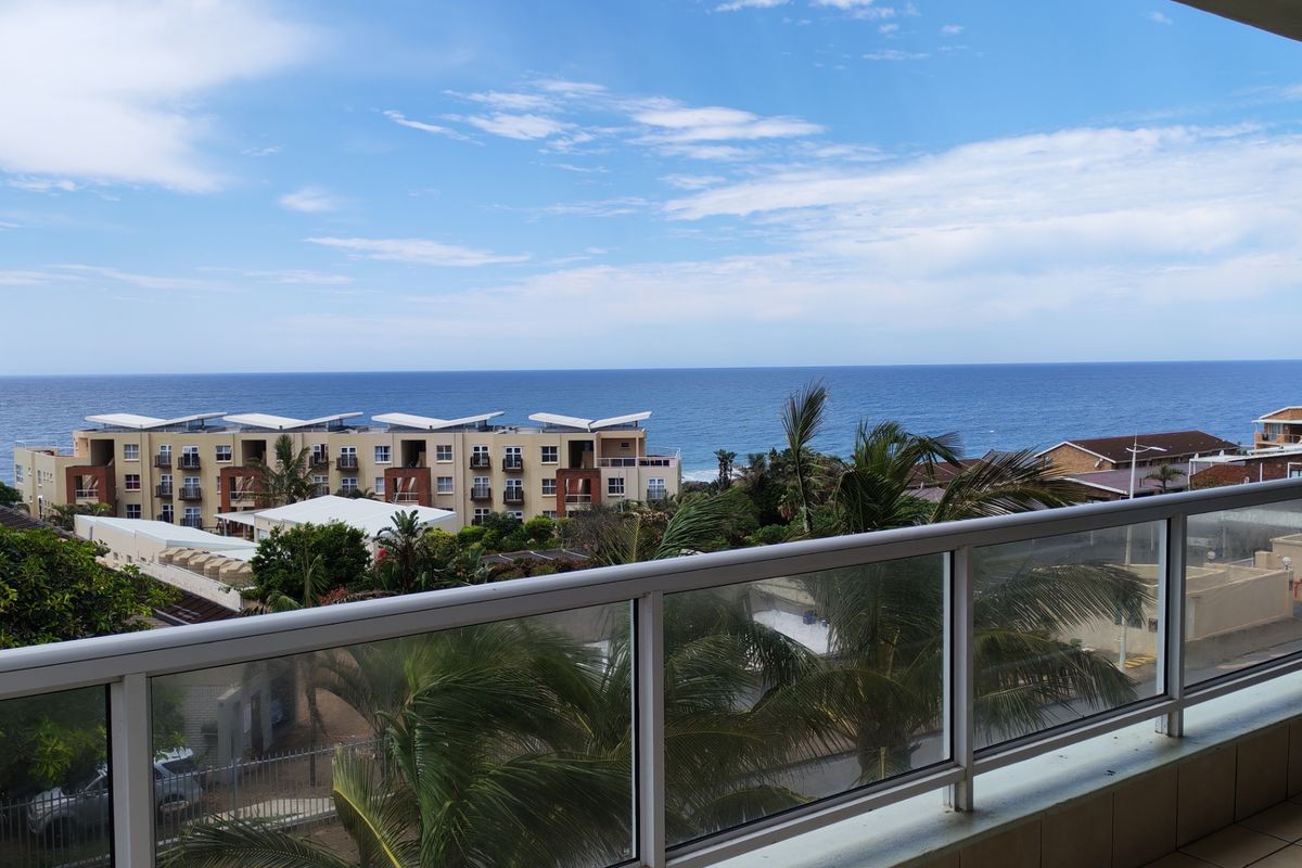 A clear elevated view of the ocean and surrounding coastal apartment complex from a private balcony with a modern glass balustrade.