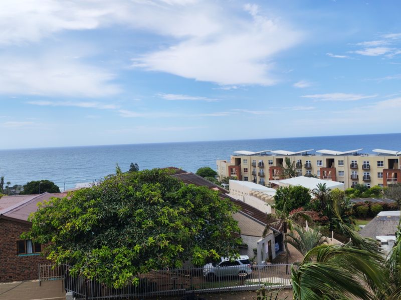 A scenic elevated view overlooking the coastline, surrounding residential properties, and lush greenery under a clear blue sky.