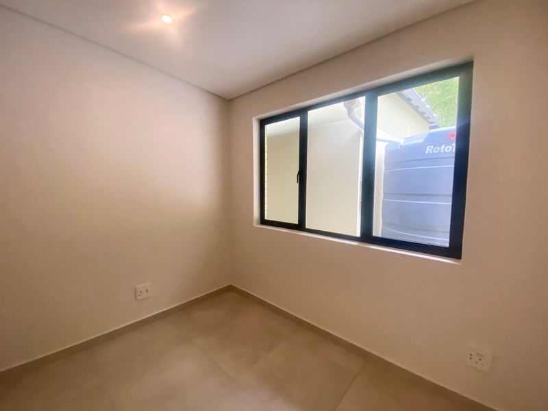 A clean, empty bedroom featuring neutral tiled flooring, recessed ceiling lighting, and modern black-framed windows overlooking the exterior side area.