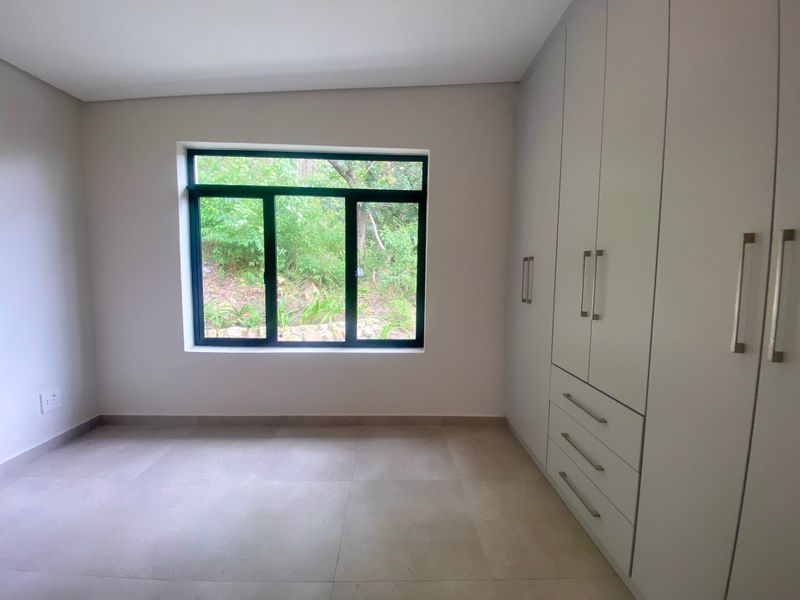 A bright, modern bedroom featuring neutral floor tiles, built-in cupboards with sleek handles, and a large window overlooking a lush garden.