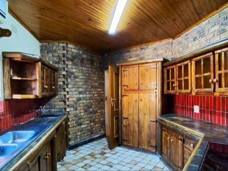 A rustic kitchen featuring exposed brick walls, timber cabinetry, tiled countertops, and a double basin sink, offering a traditional aesthetic with functional layout.