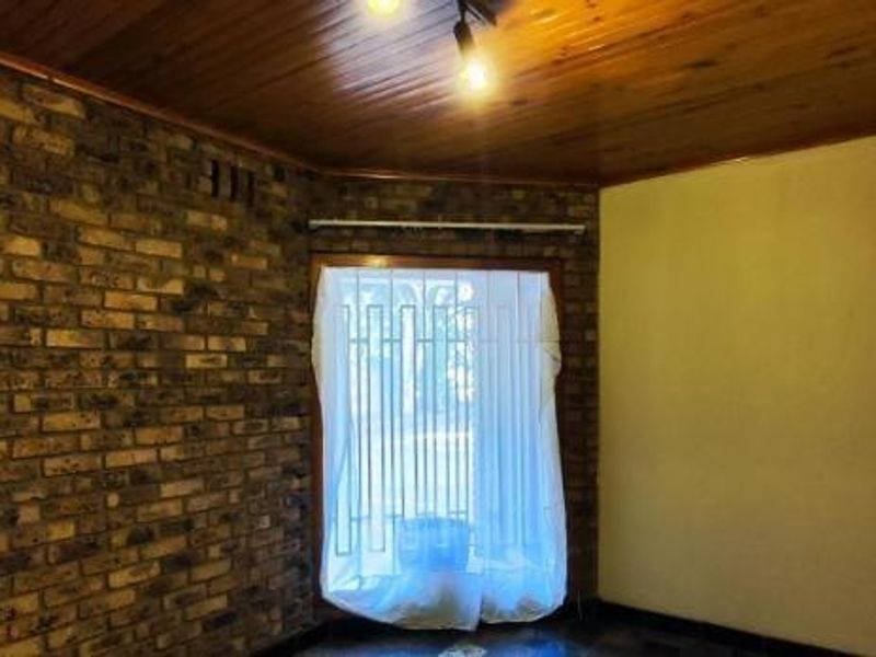 An empty living room featuring an exposed brick feature wall, timber ceiling, and a window fitted with security bars and vertical blinds.