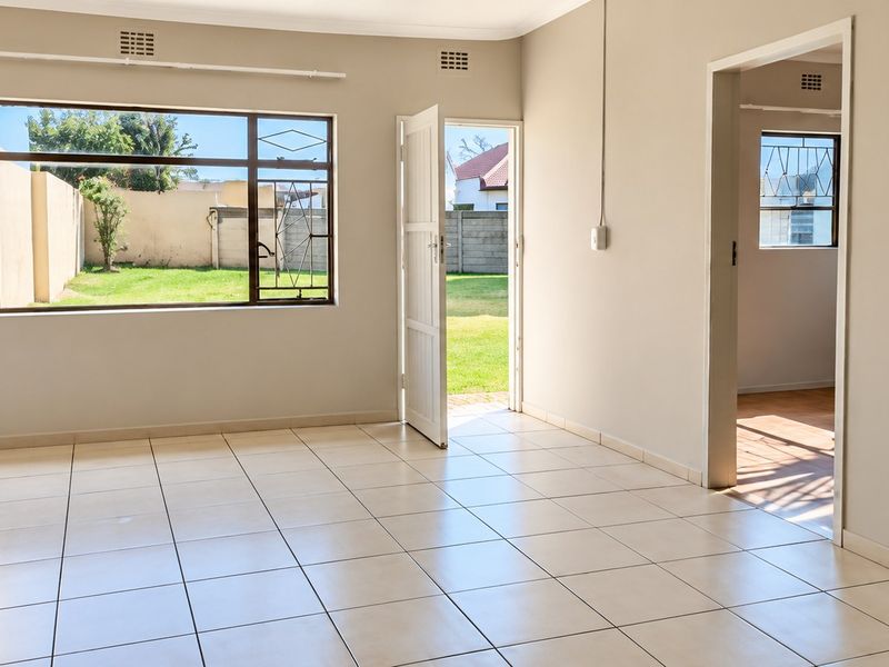 A bright, empty living area featuring neutral tiled flooring, large windows with security bars, and direct access to the outdoor garden space.