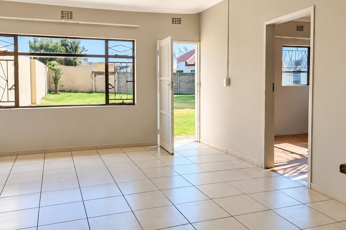 A bright, empty living area featuring neutral tiled flooring, large windows with security bars, and direct access to the outdoor garden space.