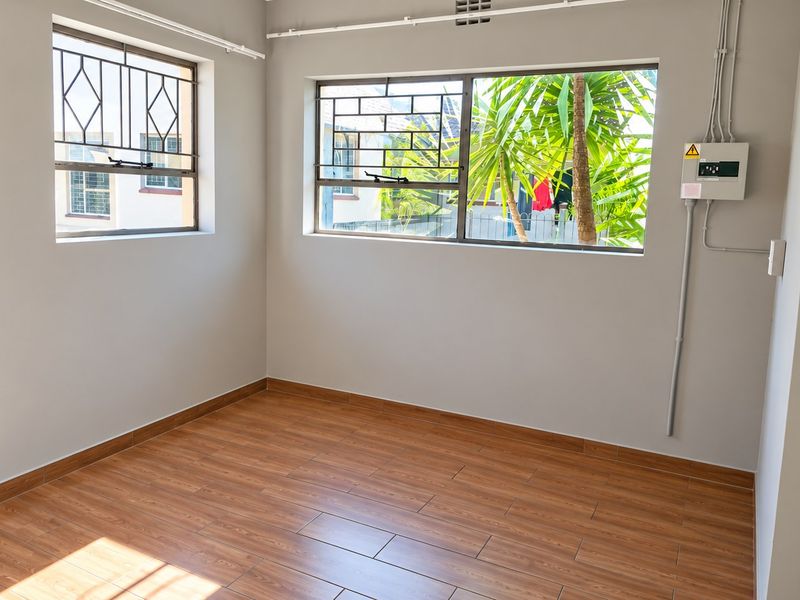 A bright, empty bedroom featuring wood-look tile flooring, neutral walls, and secure windows with fitted burglar bars.