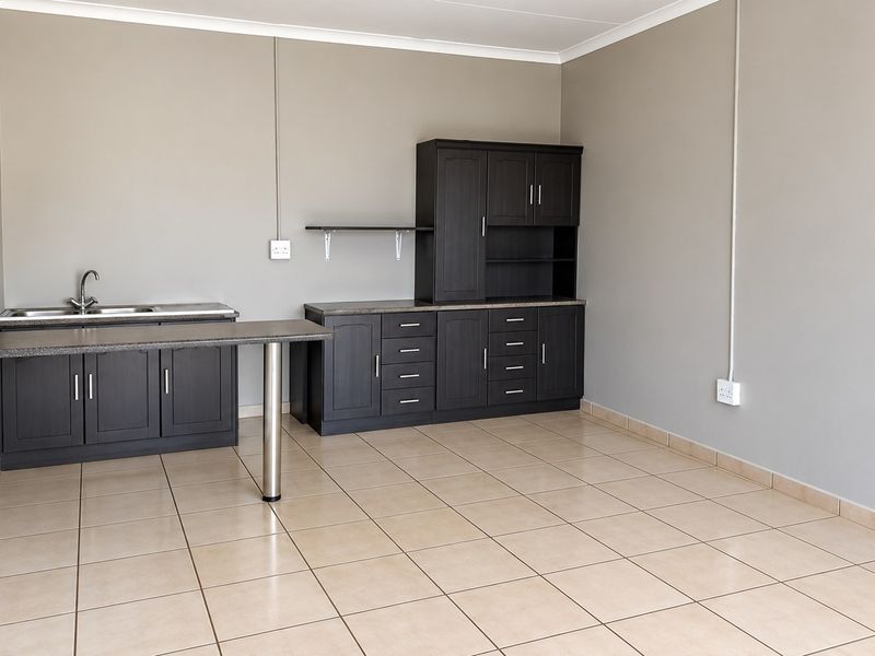 A clean, empty kitchen area featuring dark wood-look built-in cabinetry, laminate countertops, a stainless steel sink, and neutral tiled flooring.
