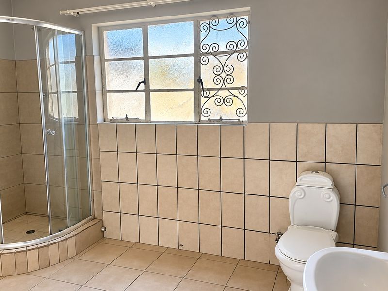 A functional bathroom featuring a corner shower enclosure, pedestal basin, toilet, and neutral ceramic tiling throughout with security bars on the window.