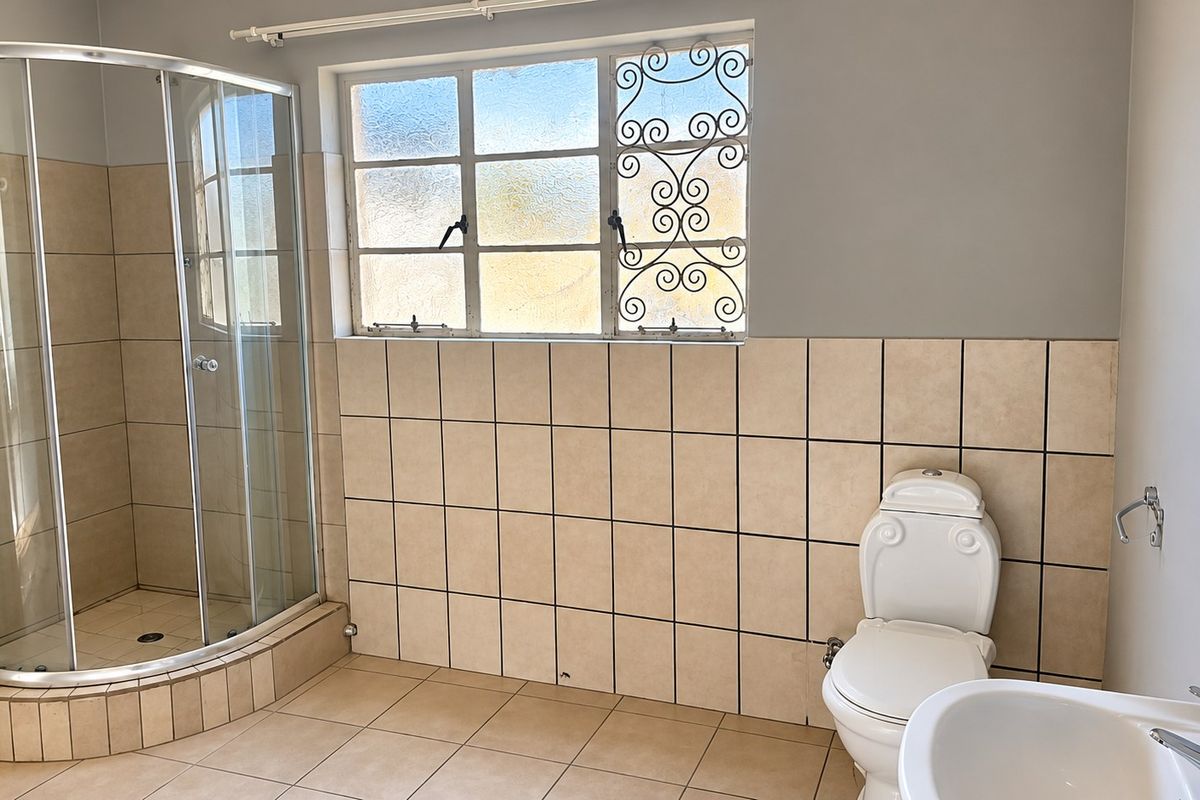 A functional bathroom featuring a corner shower enclosure, pedestal basin, toilet, and neutral ceramic tiling throughout with security bars on the window.