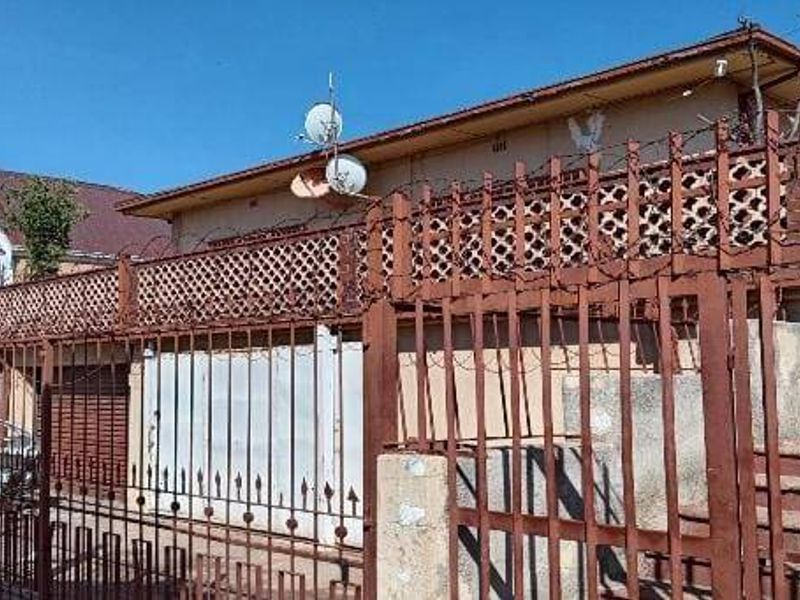 Front elevation of a residential house featuring extensive metal security fencing, barbed wire, and concrete stairs leading to the property entrance.