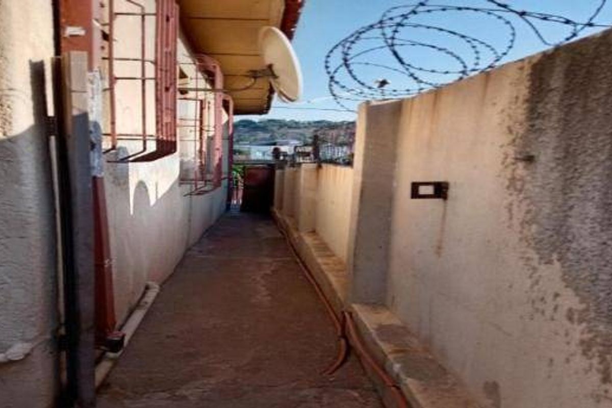 A narrow exterior side passage featuring a concrete walkway, security bars on windows, and a boundary wall topped with razor wire.