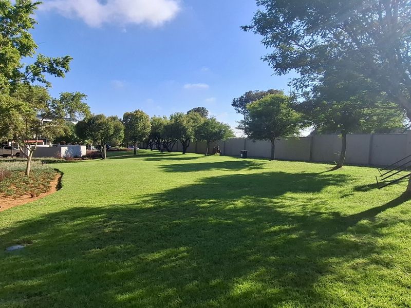 A well-maintained communal garden area featuring expansive green lawns, mature trees, and a secure perimeter wall within the apartment complex grounds.