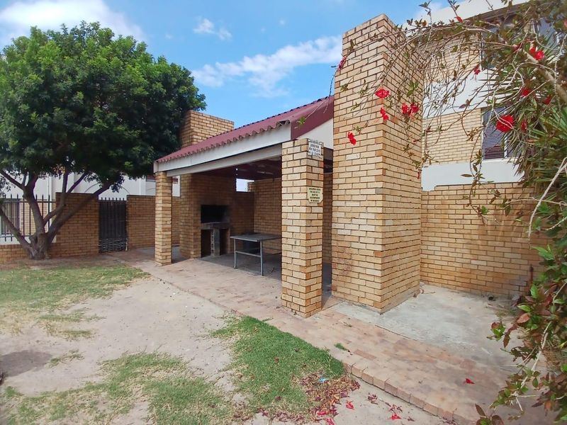 A covered outdoor brick braai area with a tiled roof, situated in a rear garden setting with paved flooring.