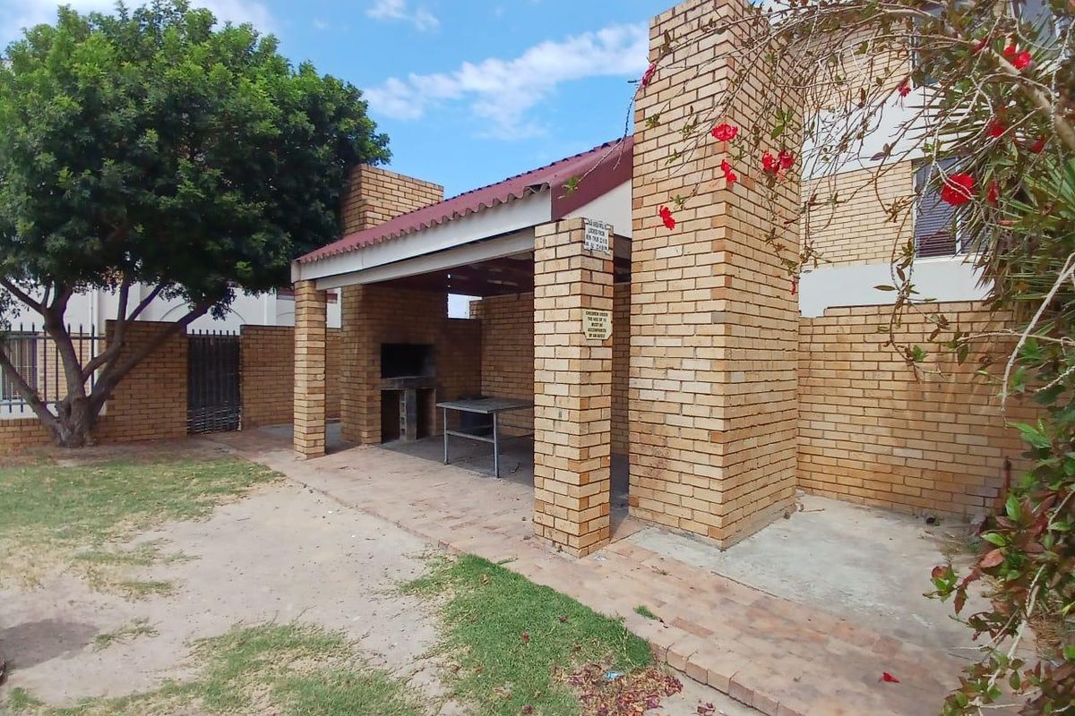 A covered outdoor brick braai area with a tiled roof, situated in a rear garden setting with paved flooring.