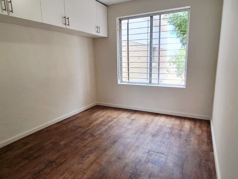 A bright, empty bedroom featuring modern laminate flooring, built-in overhead storage cabinetry, and a window fitted with security bars.