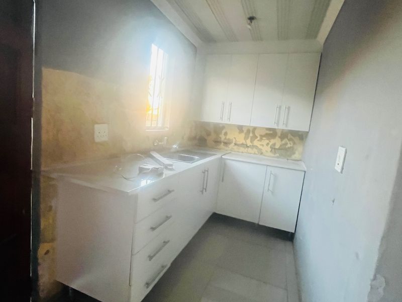 A compact kitchen area featuring white built-in cabinetry and a stainless steel sink, currently requiring wall finishing and cosmetic updates.