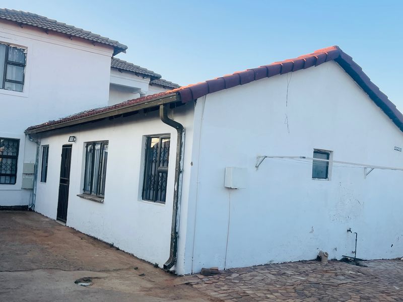 Rear view of the property showing white exterior walls, tiled roofing, security bars on windows, and a paved outdoor area.