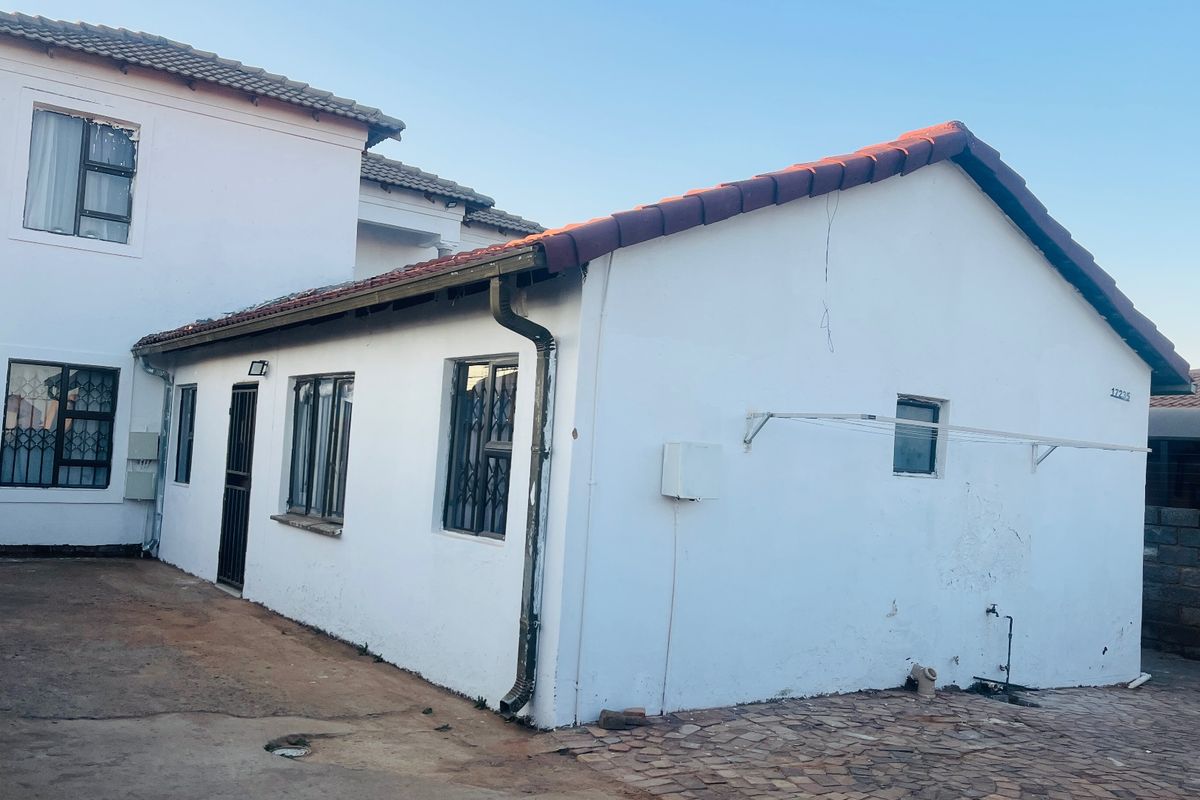 Rear view of the property showing white exterior walls, tiled roofing, security bars on windows, and a paved outdoor area.