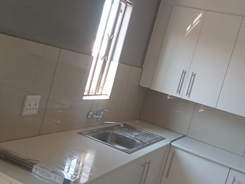 A compact kitchen area featuring white built-in cabinetry, a stainless steel sink, and tiled splashbacks, currently requiring final wall finishing.