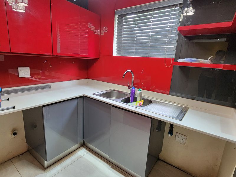 A compact kitchen featuring vibrant red glass splashbacks, grey cabinetry, white stone countertops, and a stainless steel double sink with a modern mixer tap.