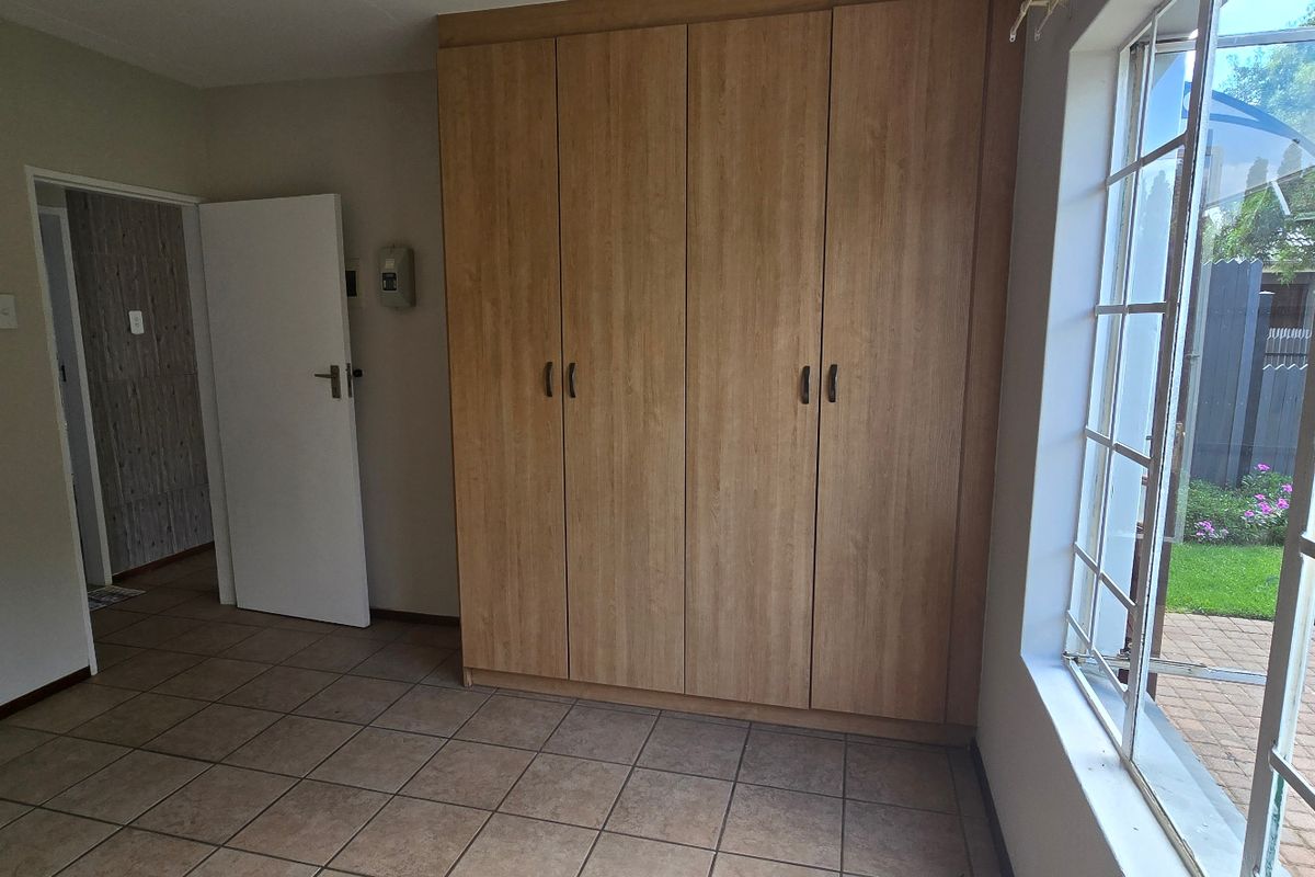 A bright, empty bedroom featuring built-in wooden cupboards, tiled flooring, and large windows with security bars overlooking the garden.