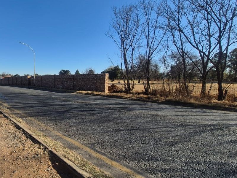 A street-level view showing an asphalt road, concrete curbing, and a brick boundary wall bordering an open field.