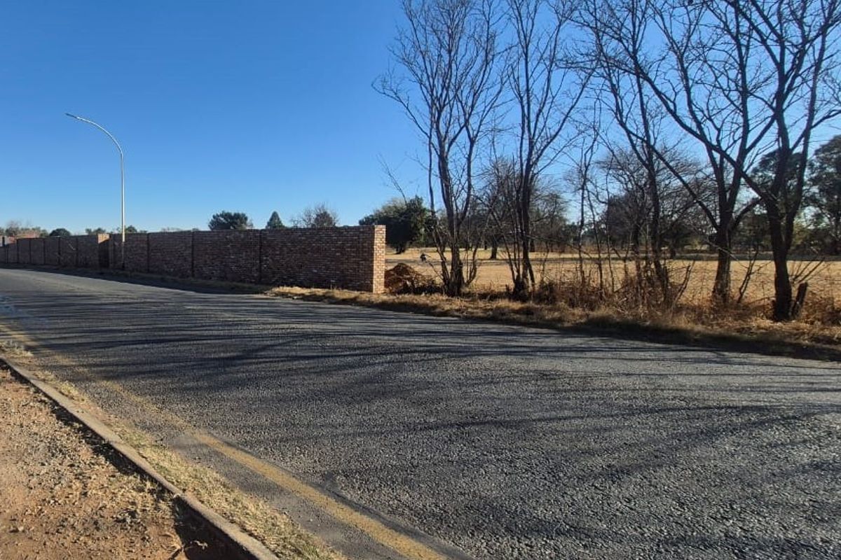 A street-level view showing an asphalt road, concrete curbing, and a brick boundary wall bordering an open field.