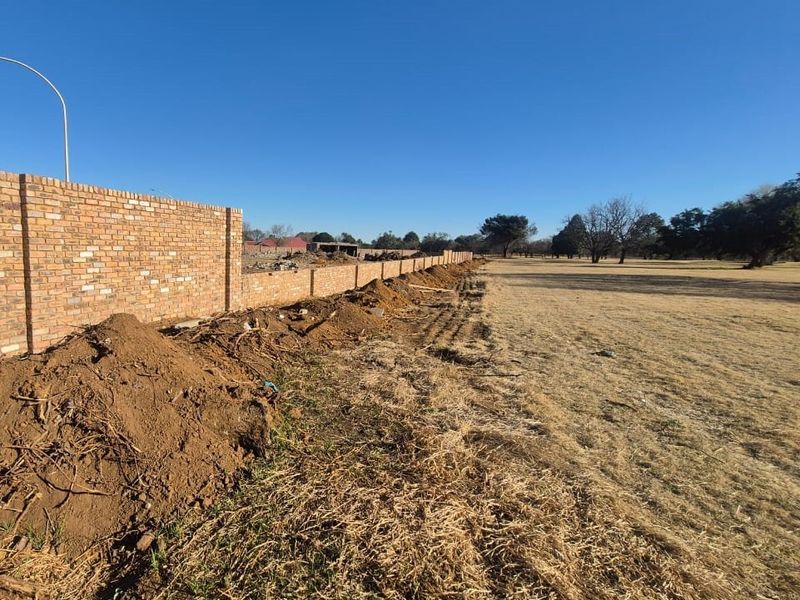 A view of a long brick boundary wall bordering an open, unlandscaped field with disturbed soil along the perimeter.