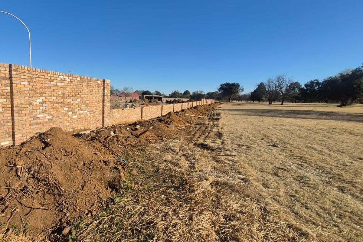 A view of a long brick boundary wall bordering an open, unlandscaped field with disturbed soil along the perimeter.