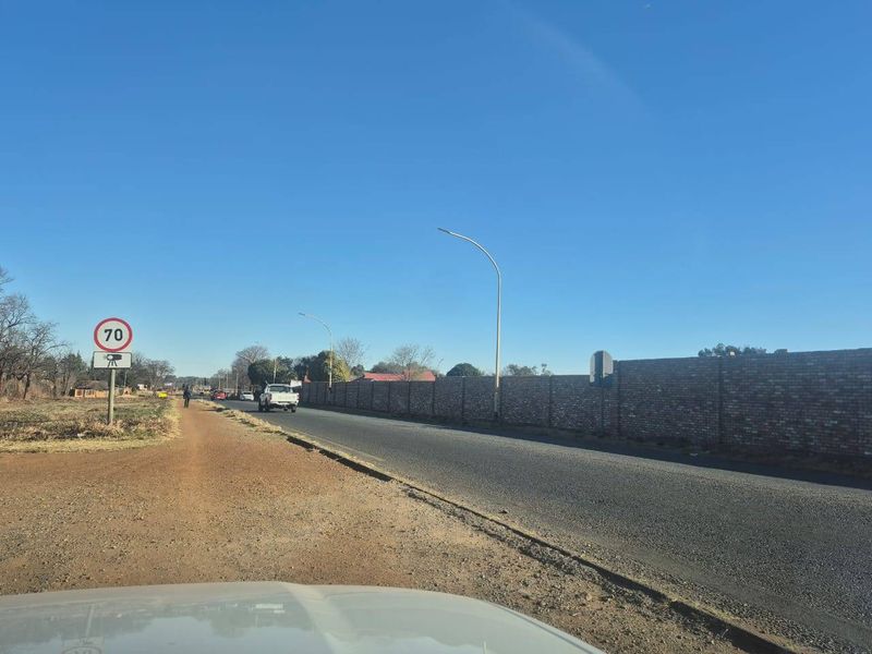 A street-level view showing the asphalt road, roadside verge, and a long brick boundary wall bordering the property.