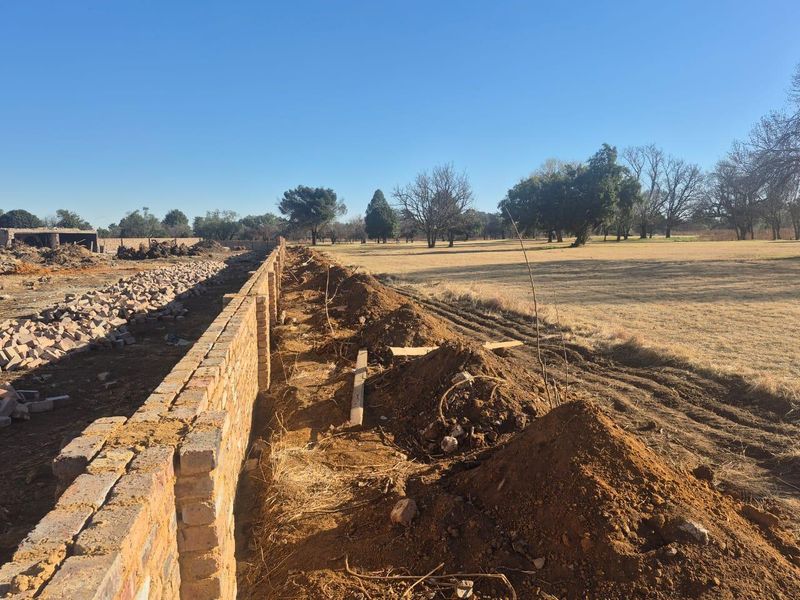 An exterior view of a commercial property site showing an ongoing brick boundary wall construction project with cleared land and surrounding landscape.