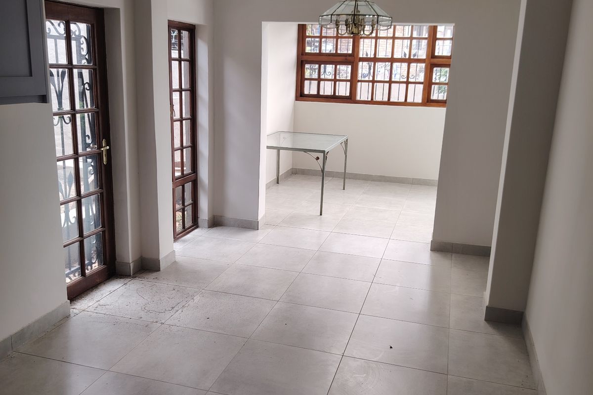 An open-plan living area featuring neutral tiled flooring, decorative ceiling fixtures, and wooden-framed windows and doors, offering a bright and versatile space.