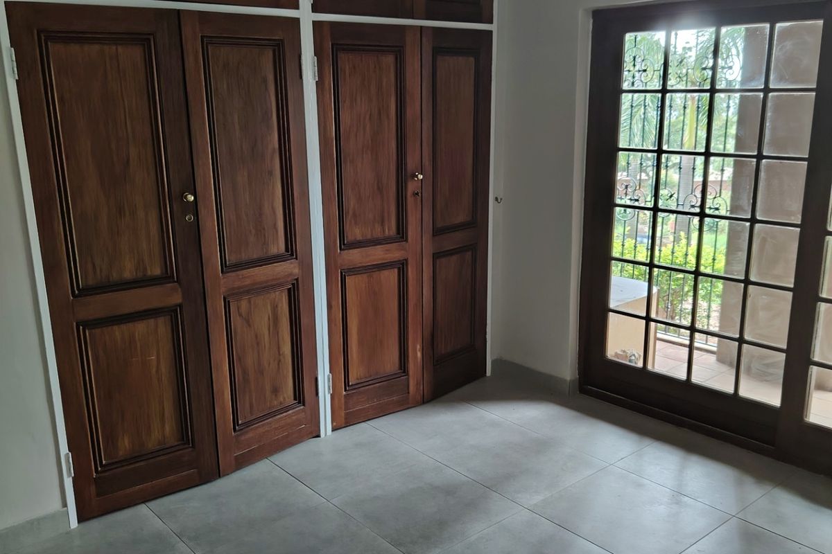 A bright, empty bedroom featuring built-in wooden cupboards, large format floor tiles, and a glass-paneled door providing access to the exterior.