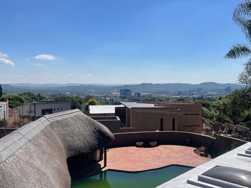 Elevated view overlooking a swimming pool, thatched roof structure, and terracotta patio with expansive urban landscape in the background.