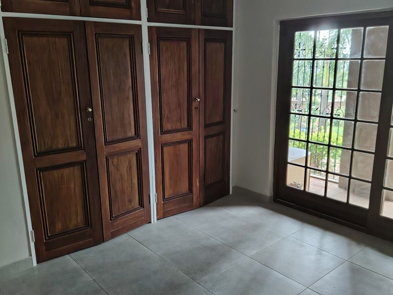 A bright, empty bedroom featuring built-in wooden cupboards, large format floor tiles, and a glass-paneled door providing access to the exterior.