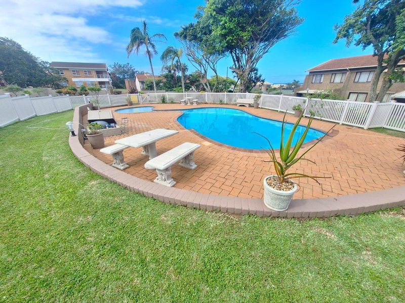 A well-maintained communal swimming pool area featuring a paved deck, surrounding lawn, and secure white perimeter fencing.