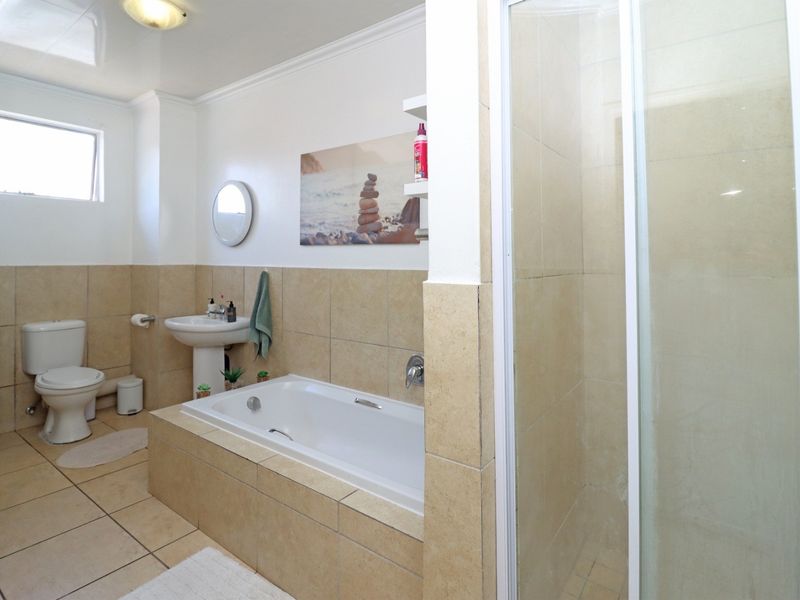 A functional bathroom featuring a tiled bathtub, separate glass-enclosed shower, pedestal basin, and toilet with neutral wall and floor tiling.