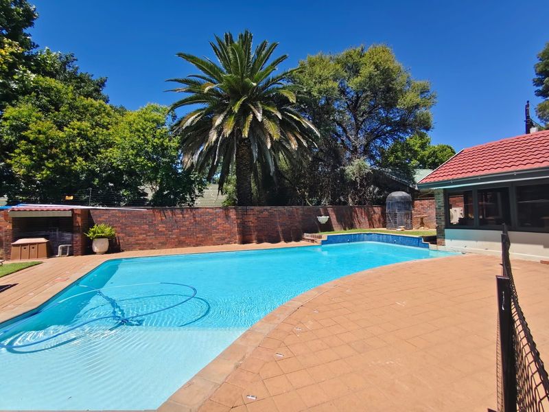 A well-maintained outdoor pool area featuring a tiled deck, brick boundary wall, and established garden surroundings.