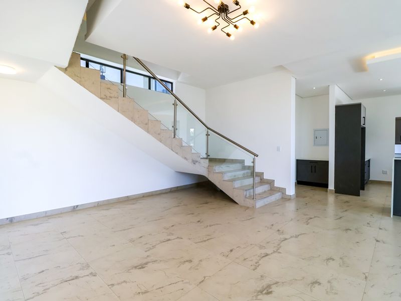 Spacious open-plan living area featuring a modern staircase with glass balustrade, marble-look tiled flooring, and a sleek kitchen with built-in cabinetry.