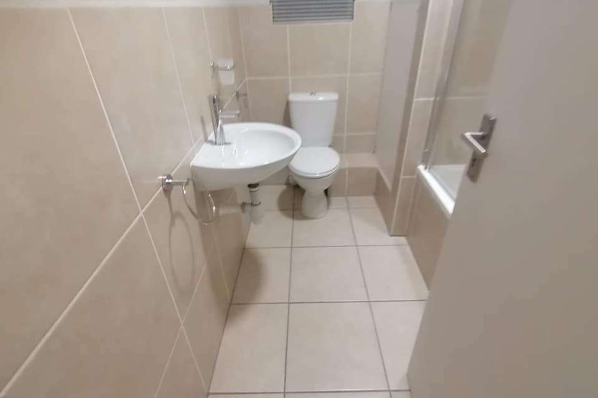 A clean, functional bathroom featuring neutral wall and floor tiles, a wall-mounted basin, toilet, and a bathtub with a glass shower screen.