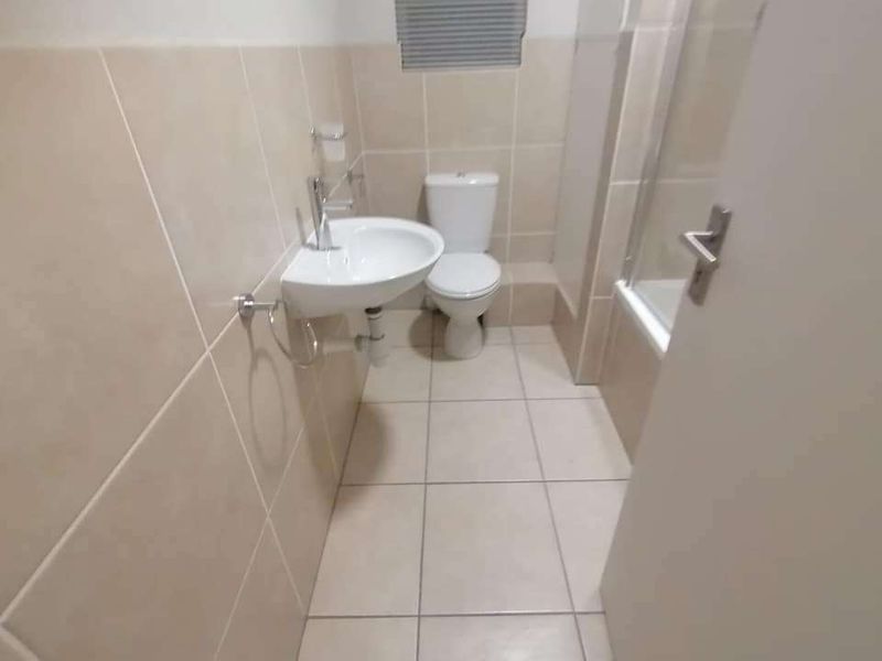 A clean, functional bathroom featuring neutral wall and floor tiles, a wall-mounted basin, toilet, and a bathtub with a glass shower screen.