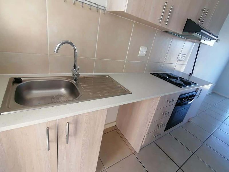A clean, functional kitchen featuring light wood-finish cabinetry, a stainless steel sink, built-in oven, electric hob, and neutral tiled backsplash and flooring.